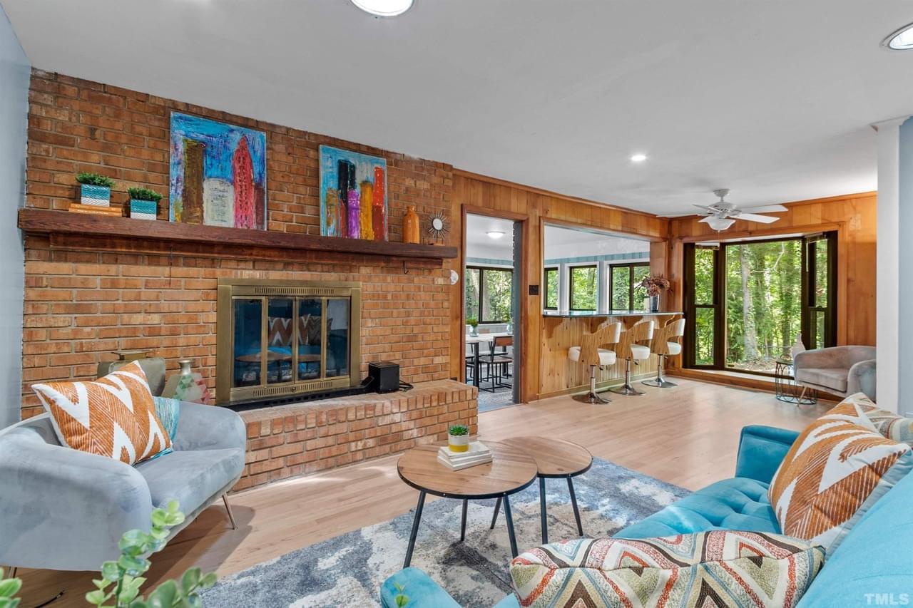 Large living room with light wood floor, wood paneled walls, brick fireplace, brightly colored art, glass doors, view to wooded lot